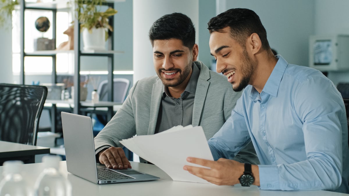 Two male colleagues review health insurance offerings on a laptop in an office. 