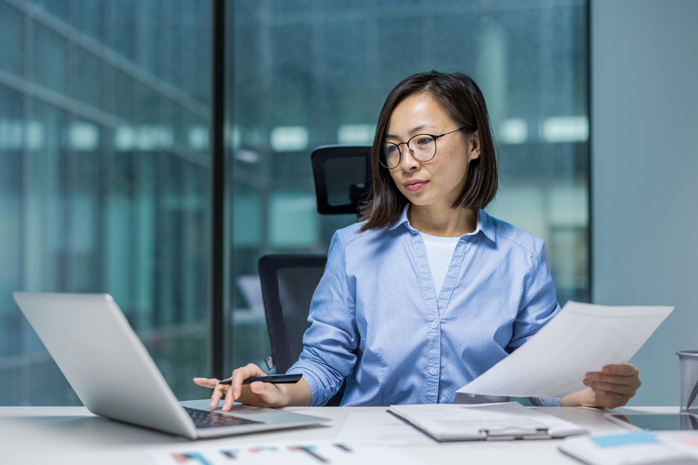 Female businesswoman engaged in work at her desk, using a laptop and reviewing documents. 